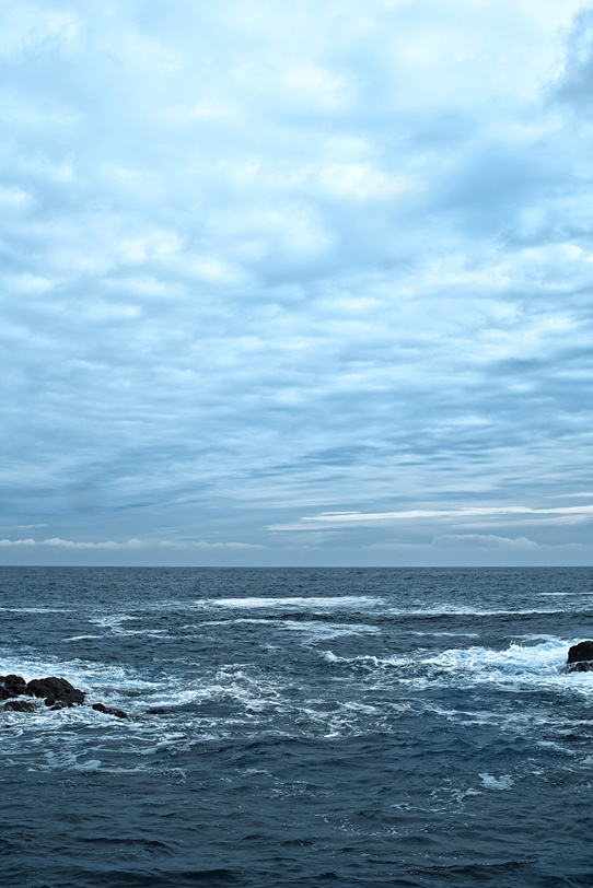 薄曇りの空と岩に砕ける波の写真画像